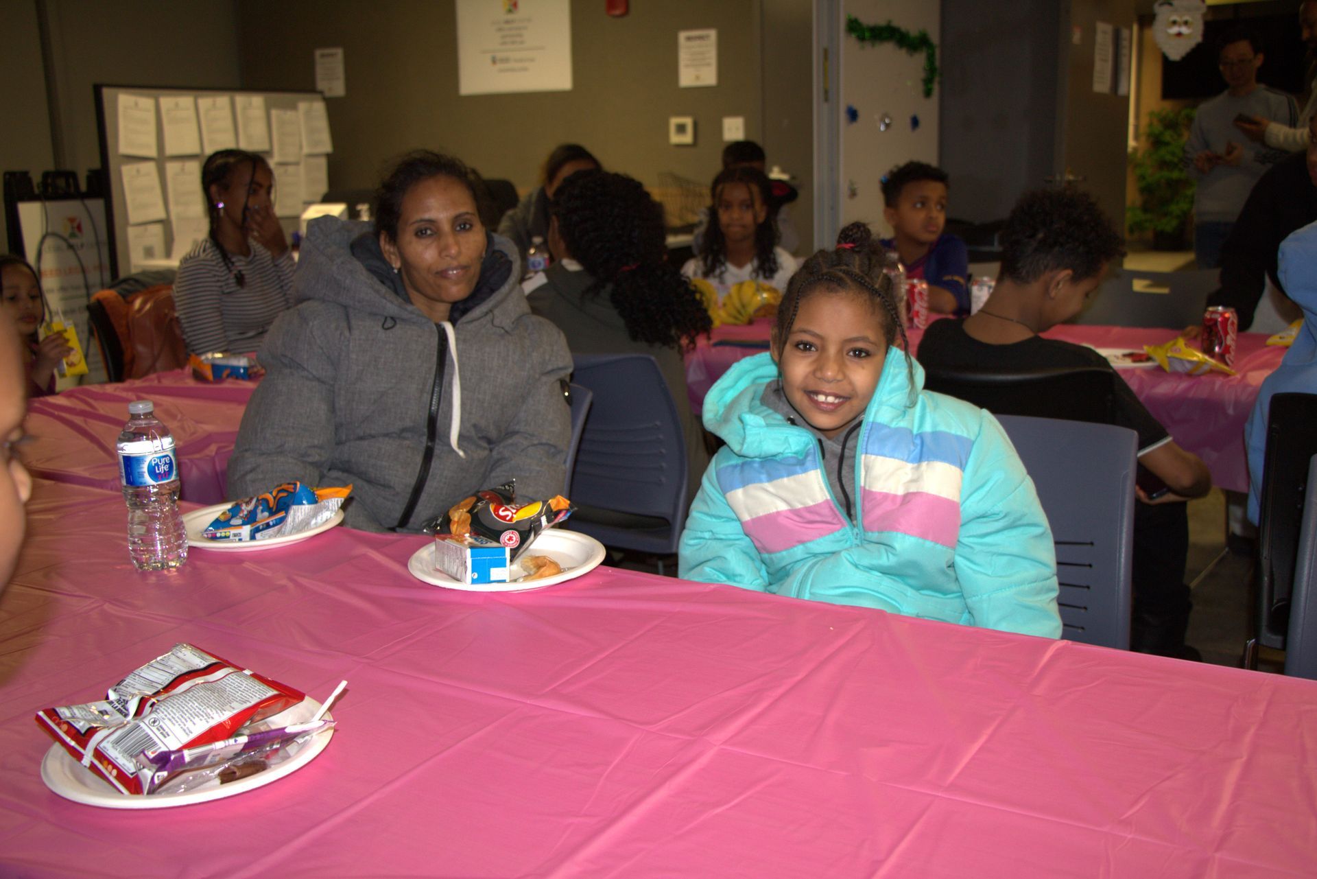 Two people smiling at a pink table, treats and drinks, other people in the background.