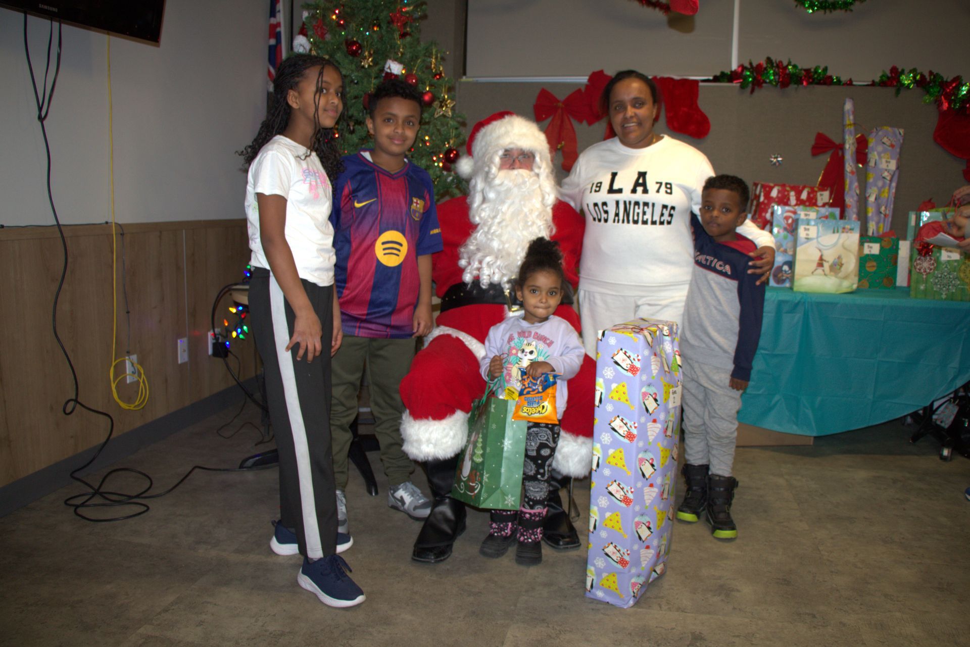 Family with Santa at a Christmas event, posing with wrapped gifts and tree in background.
