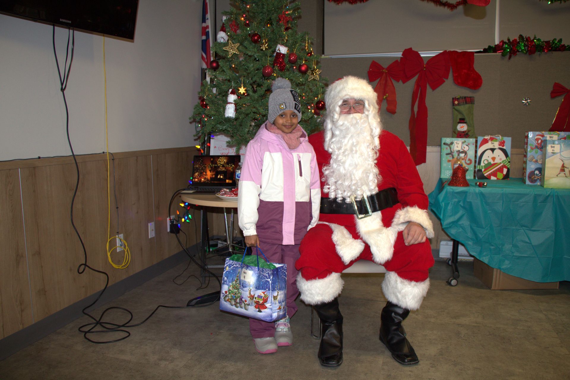 Girl with gift poses next to Santa Claus by a Christmas tree; indoors with decorations.