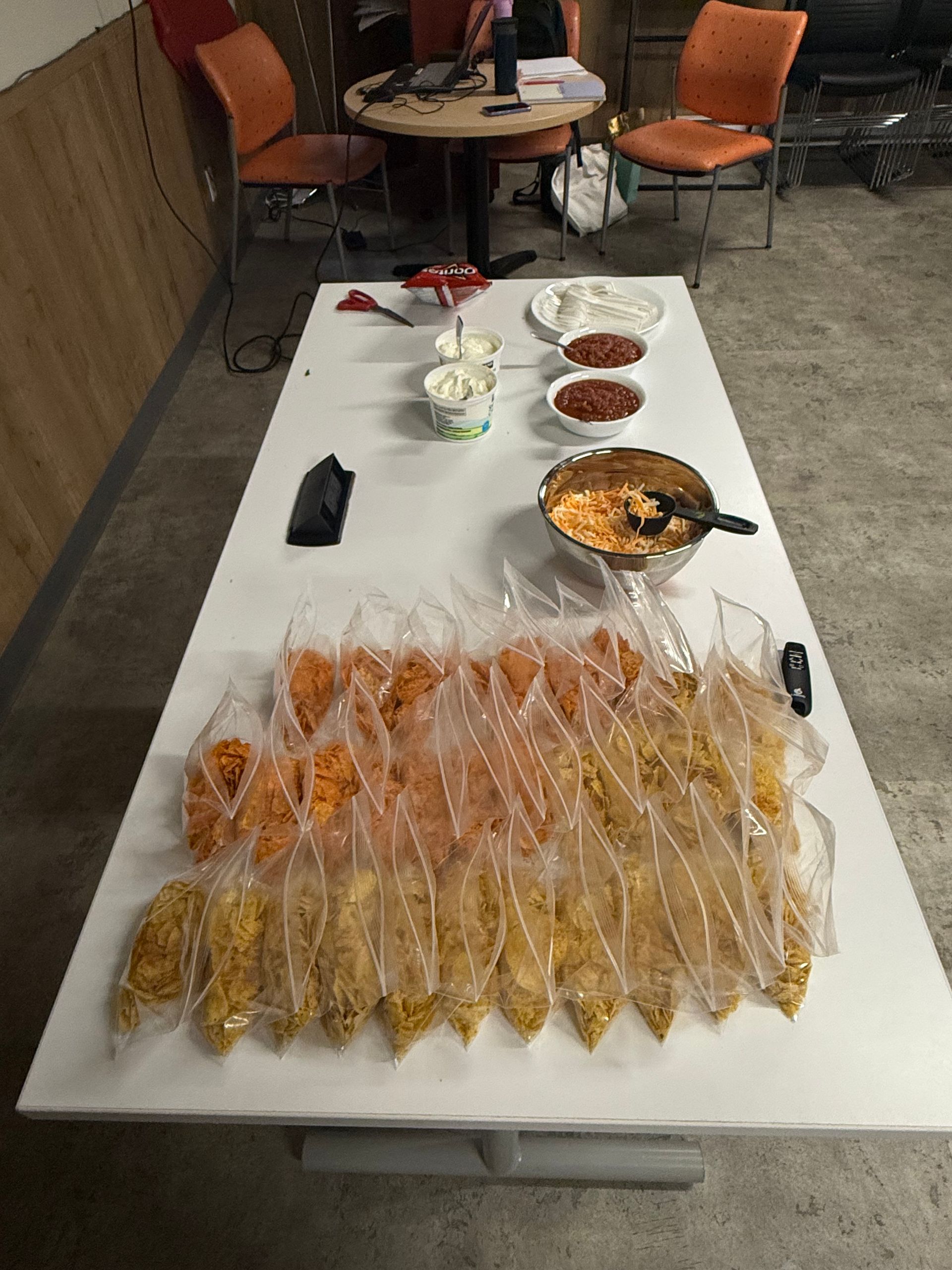 A table with many bags of chips, dips, and a bowl of queso, presumably for a gathering.