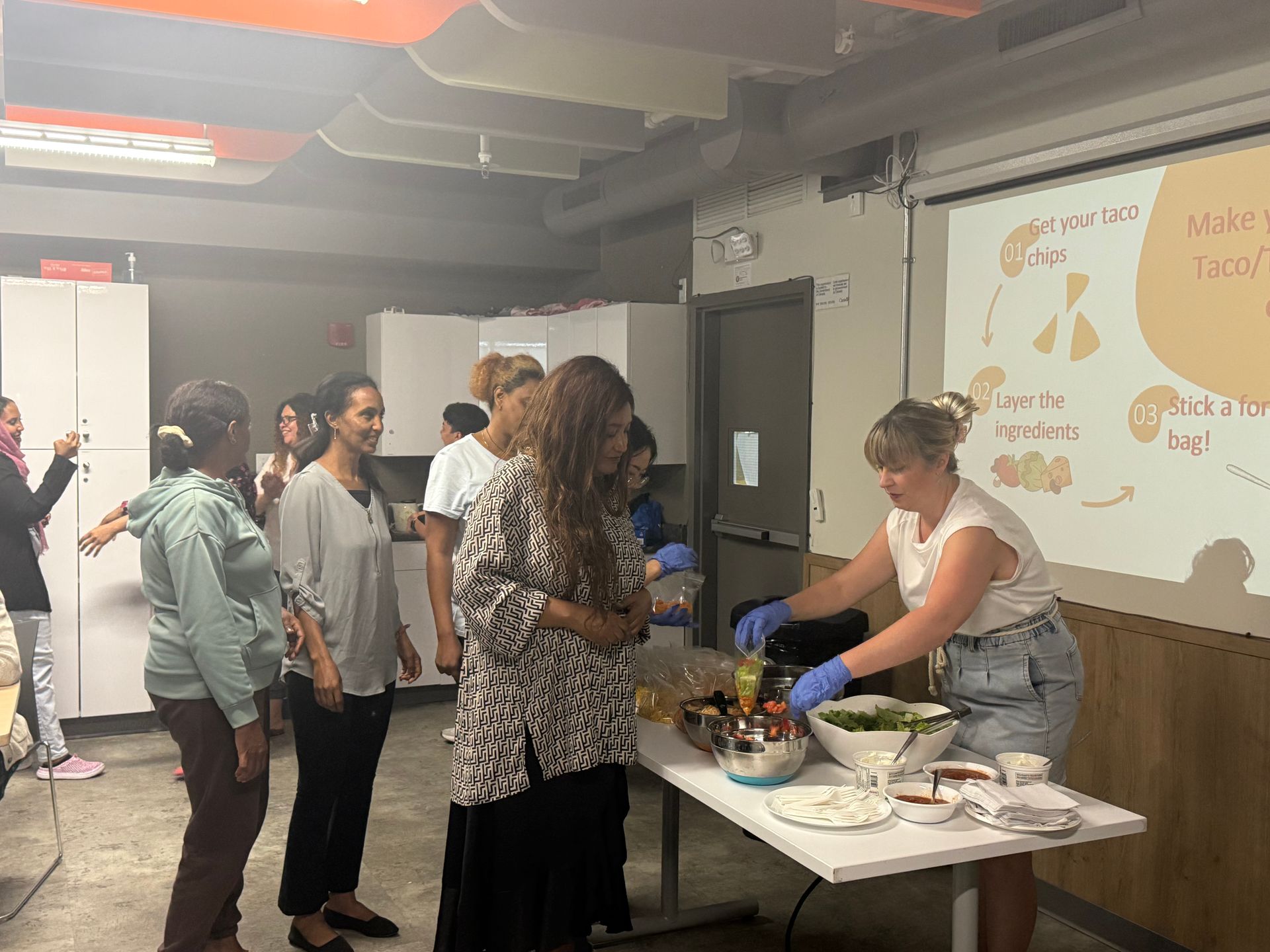A woman prepares food on a table as others watch. A projector screen displays text and diagrams.