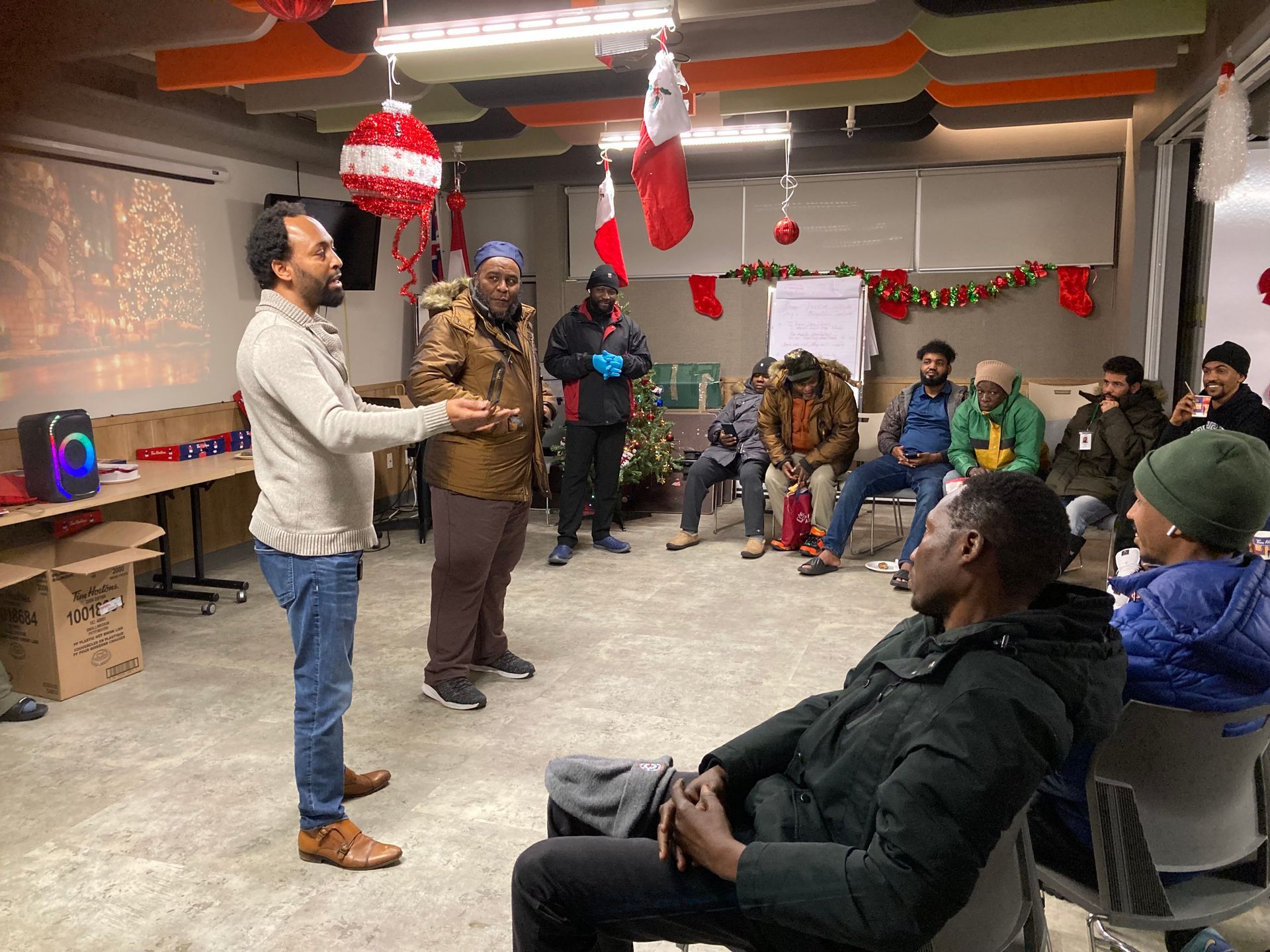 Group of people in a decorated room, listening to a man speaking. Holiday decorations.