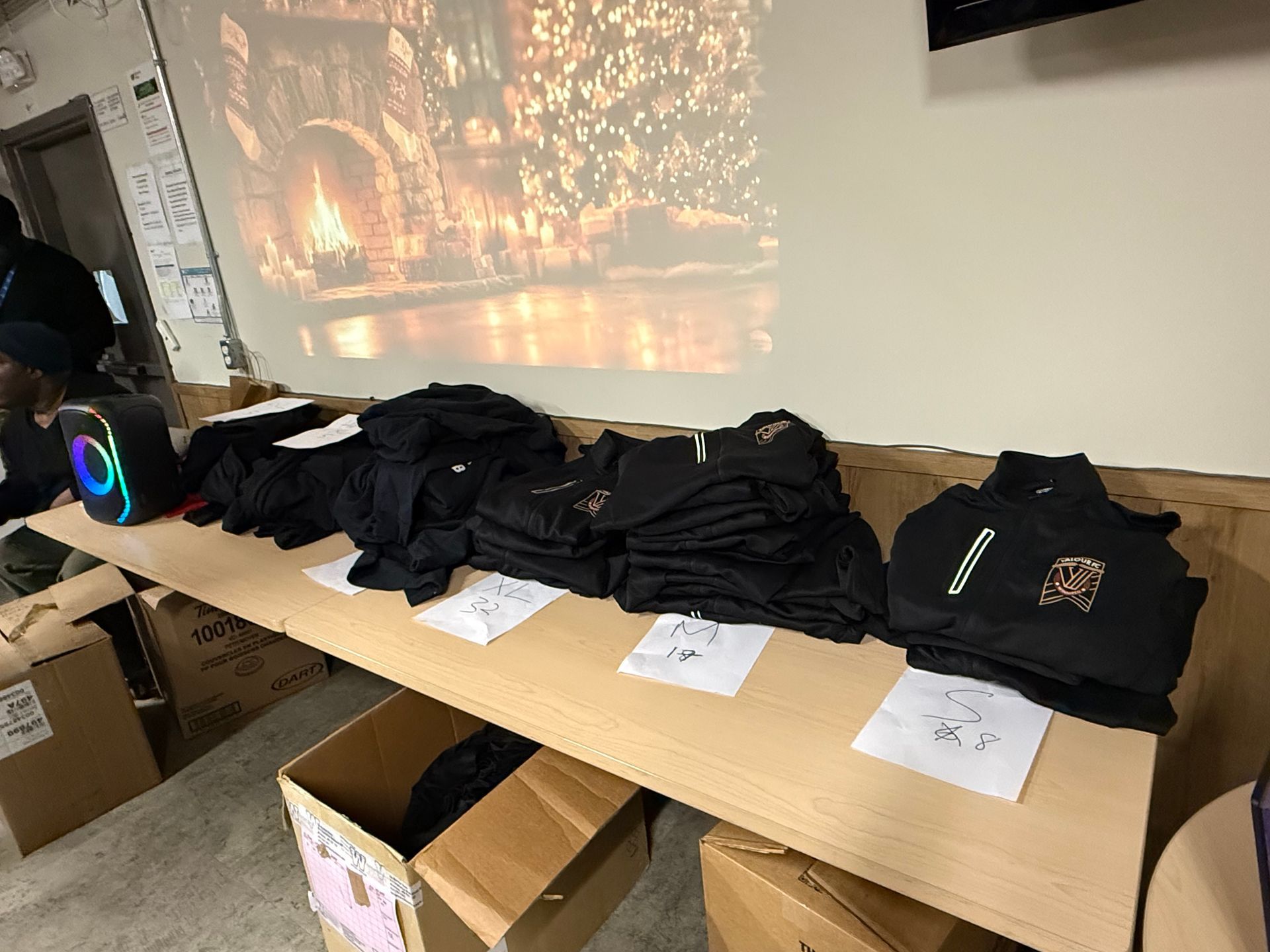Table displaying stacks of black clothing items, a projector screen with a warm scene, boxes, and a speaker.