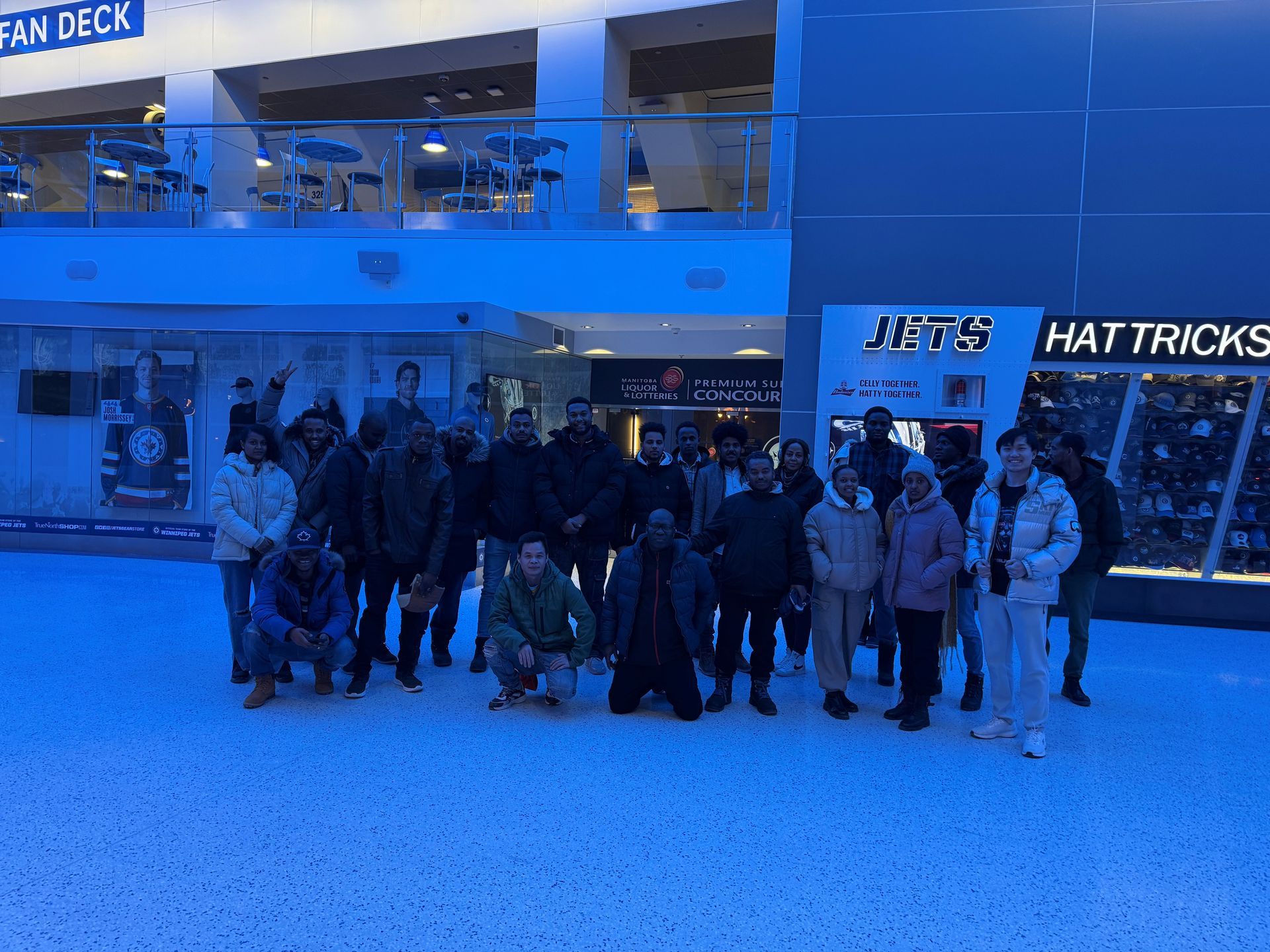 Group of people pose in front of a Jets Hat Tricks store. Indoor, blue-lit setting.