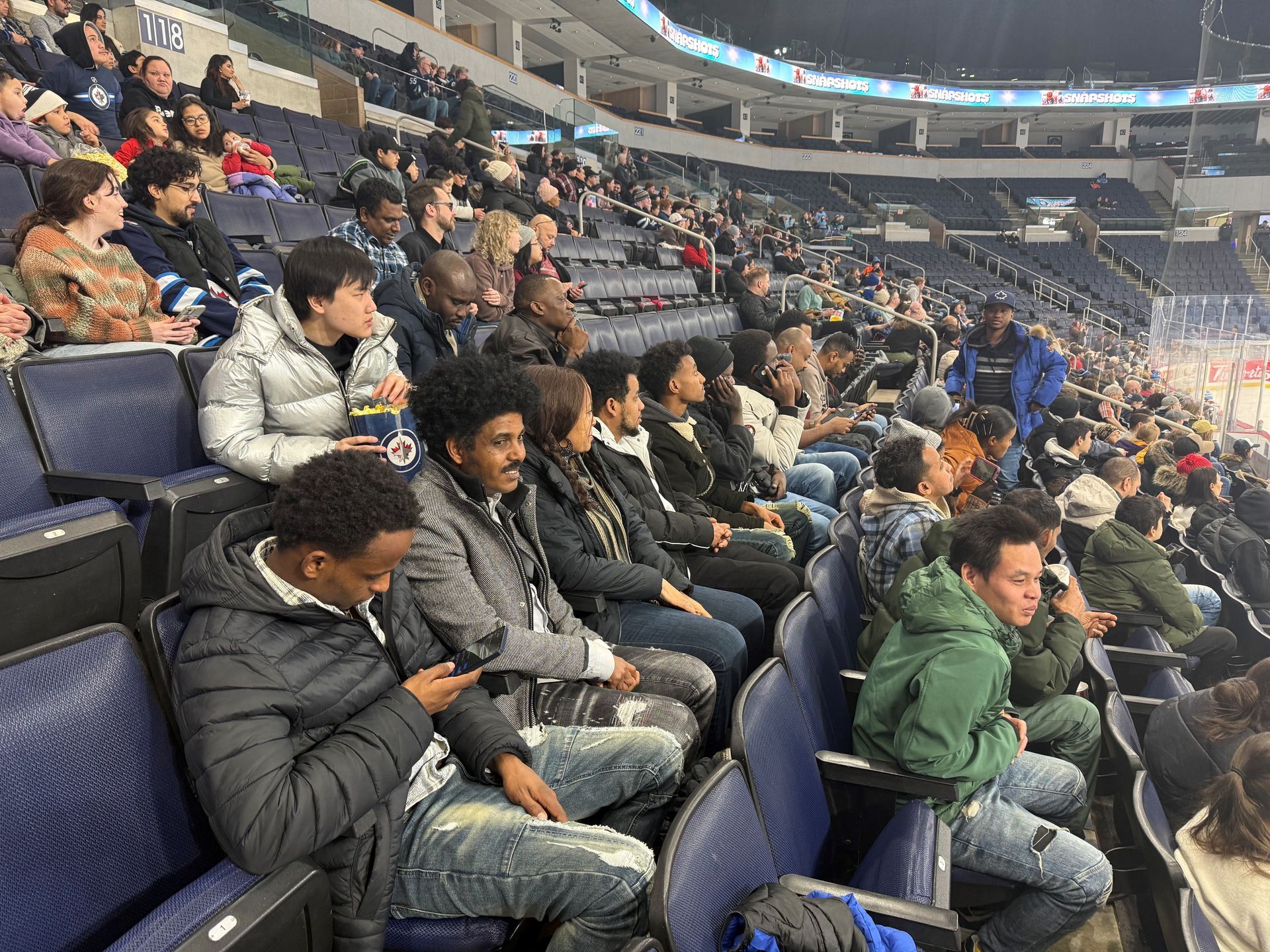 People seated in arena stands, watching an event. Blue seats, mostly dark clothing. Dim lighting.