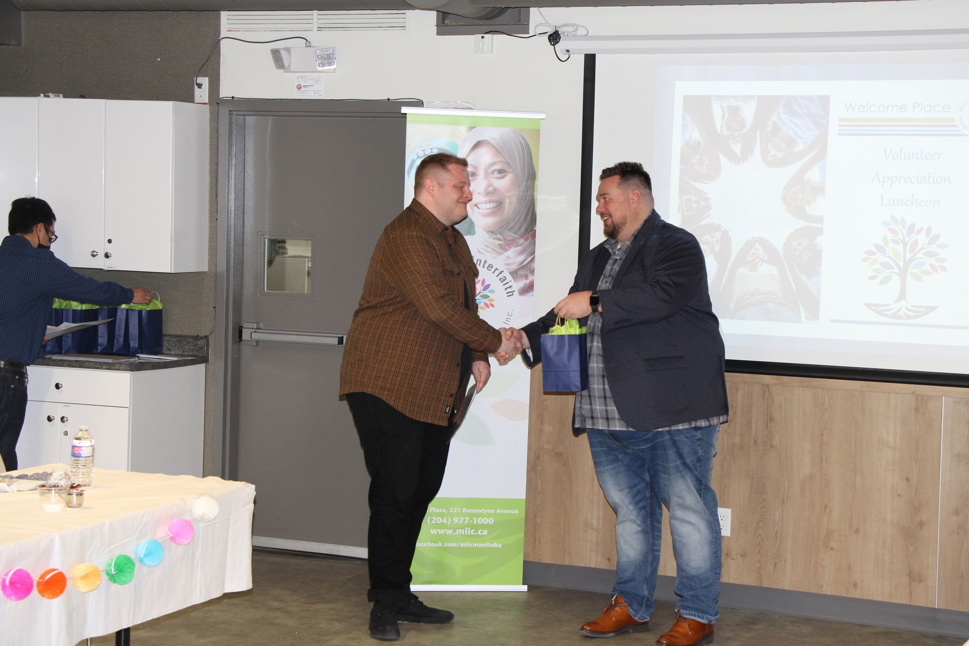 Two men are shaking hands in front of a projector screen in a room.