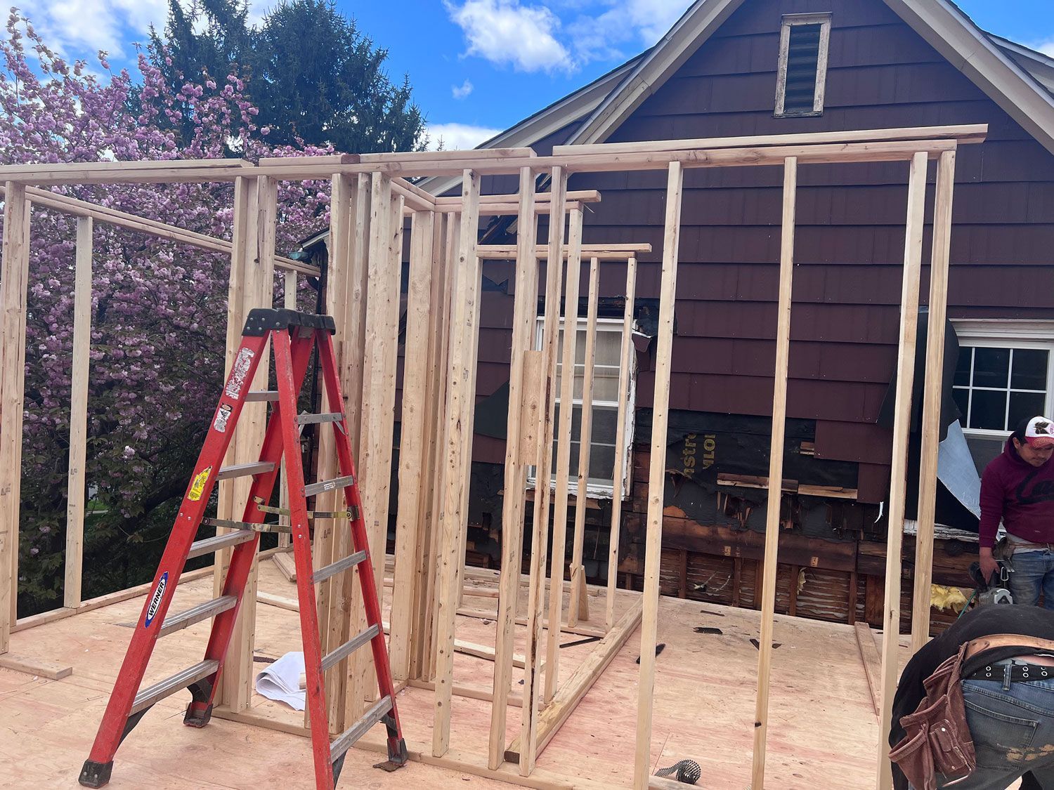 Construction of a wooden structure attached to a brown house; a red ladder is leaning against it.