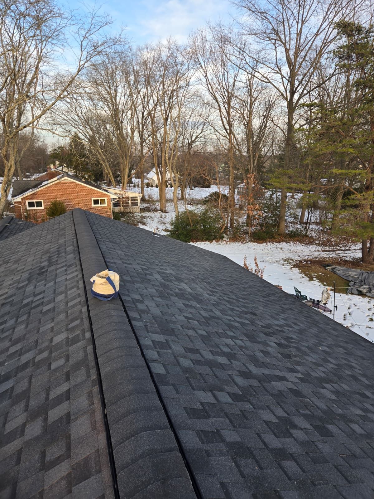 Roof shingles with a vent pipe overlooking a snowy backyard and bare trees