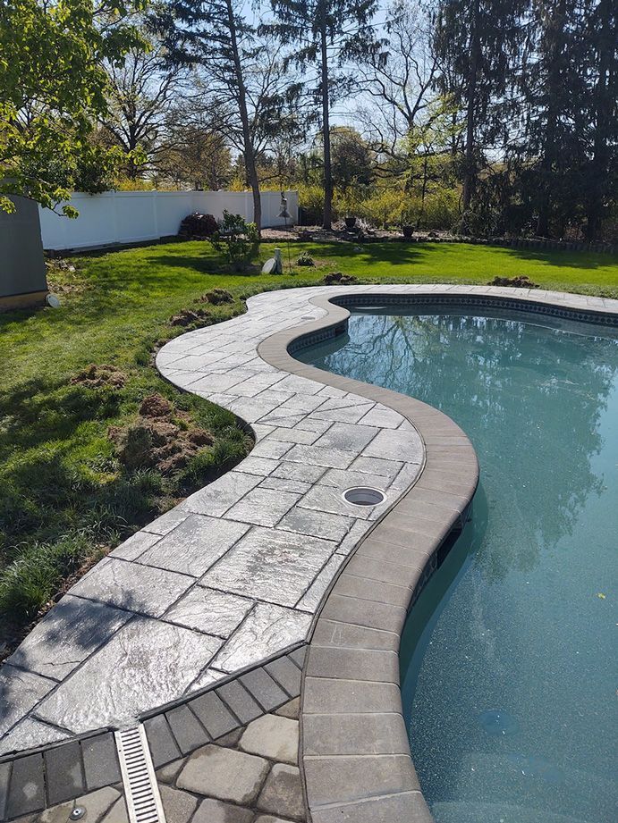 Curving stone path beside a pool, edged with brick. Green grass and trees in the background. Sunny outdoor setting.