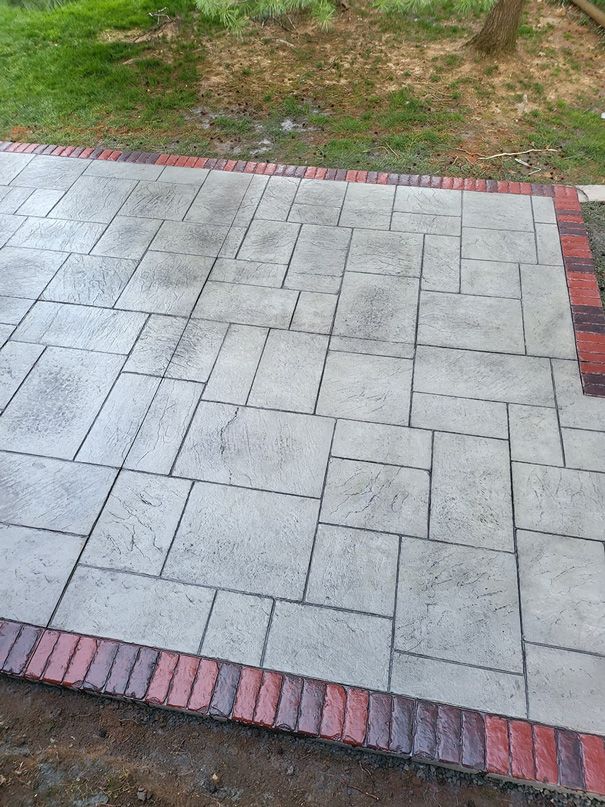 Stamped concrete patio with brick border, gray and red hues, in a grassy outdoor area.