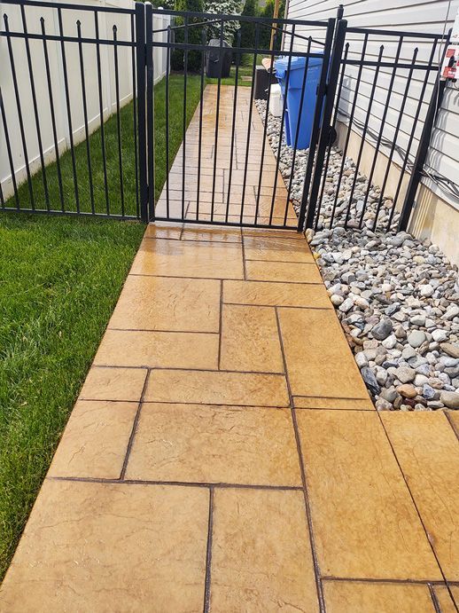 Concrete walkway with black fence, gate, and trash can; grass on left, rocks on right.