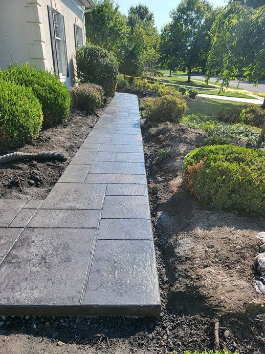 Gray stamped concrete walkway bordered by shrubs and soil in front of a house.