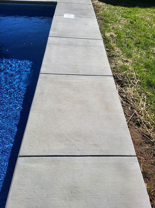Concrete pool deck next to blue water. Grass and dirt border the edge.