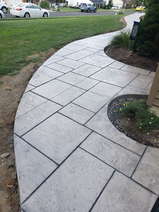Curving concrete walkway with gray rectangular tiles and dark grout, leading through a yard.