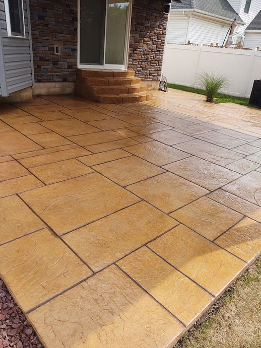 Stamped concrete patio with large square and rectangular patterns, in shades of tan and brown, near a house with steps.