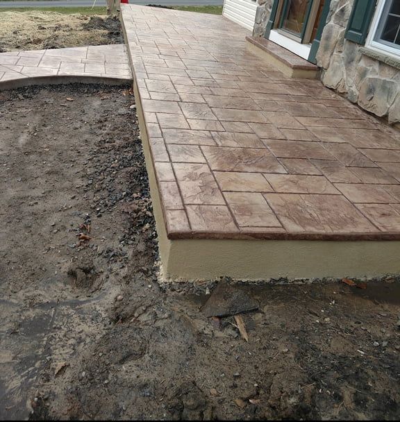 Brown, stamped concrete front porch and walkway next to a house with stone siding. Bare earth in foreground.