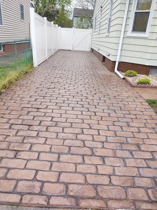 Brick-patterned driveway leading to a white gate, next to a light green house with a white fence.