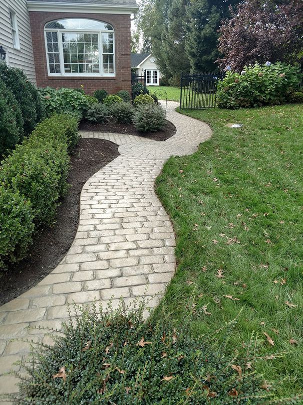 Brick pathway curves through green lawn and shrubs towards a house.