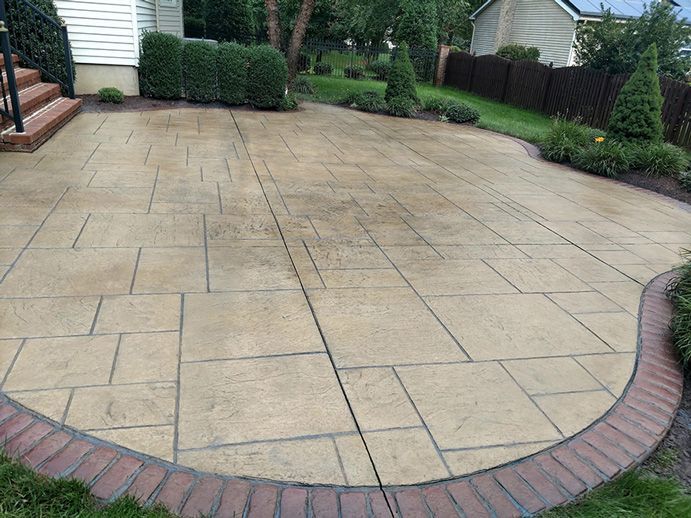 Tan stamped concrete patio with brick border, surrounded by greenery and a house in the background.