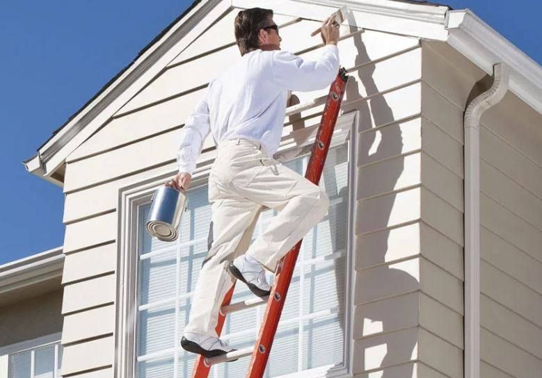 Person painting the trim of a two-story house from a ladder, holding a paint can.