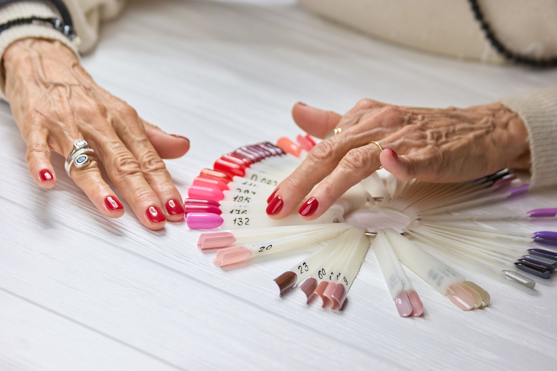 Mani con unghie dipinte di rosso, che scelgono un colore di smalto da un ventaglio di colori su una superficie bianca.