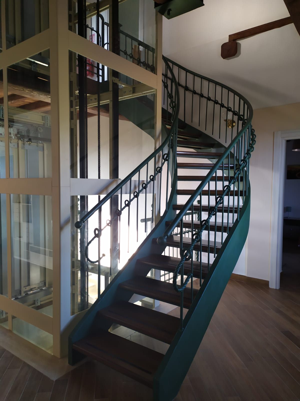 Scala a chiocciola con struttura verde, accanto a un ascensore in vetro. Pavimenti in legno e pareti bianche.