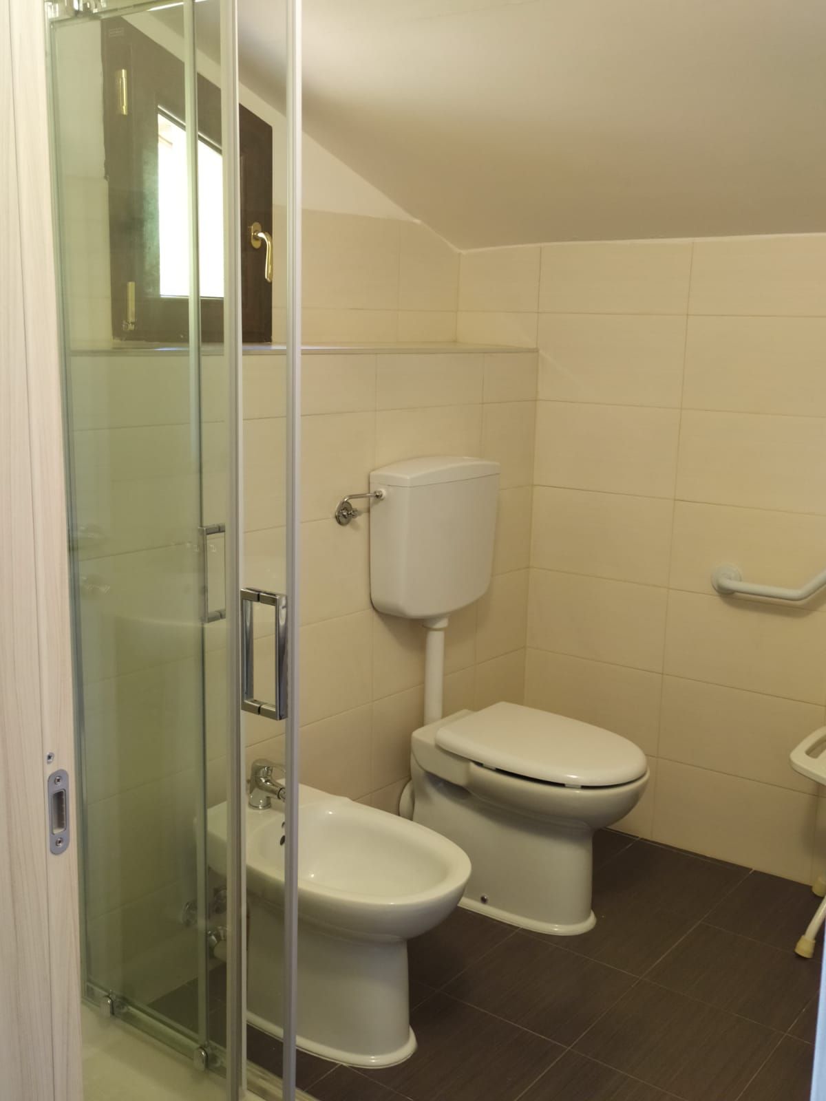 Bagno con doccia in vetro, WC e bidet. Pareti beige, pavimento marrone scuro, piccola finestra.