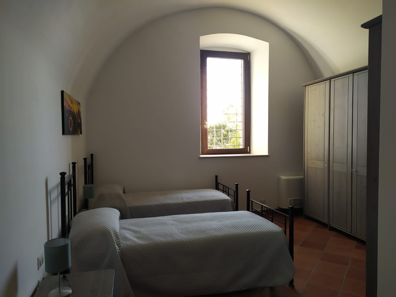 Camera da letto con due letti singoli, una finestra e un armadio. Pareti bianche e pavimento in mattoni.