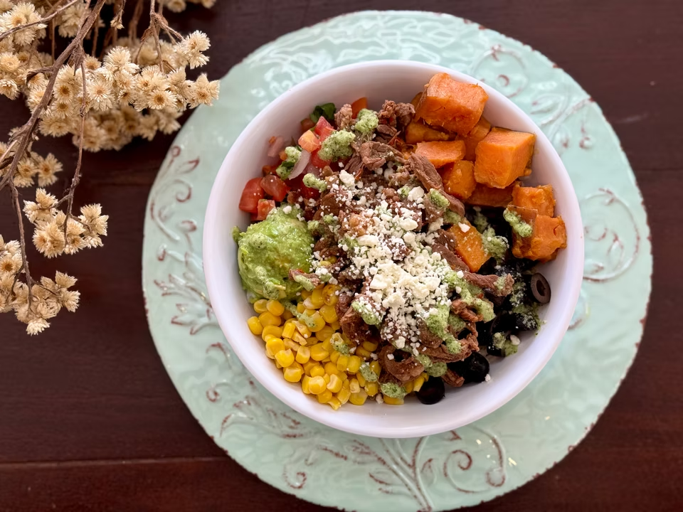Burrito bowl with sweet potato, corn, guacamole, salsa, meat, olives, and cheese.