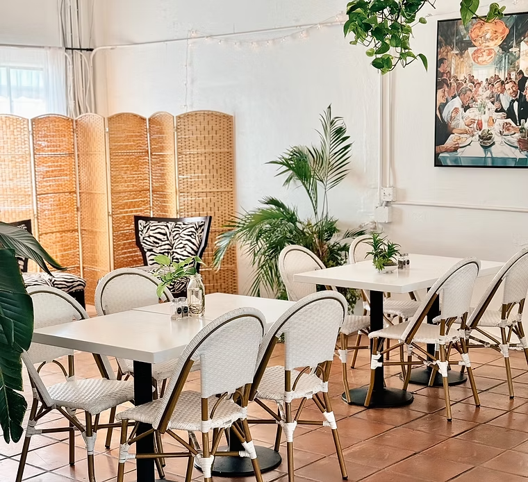 Restaurant interior with white tables, rattan chairs, plants, and a decorative screen.
