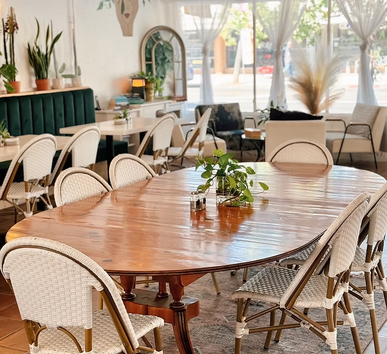 A restaurant interior with a large wooden table, white chairs, and a green booth. Natural light streams in.