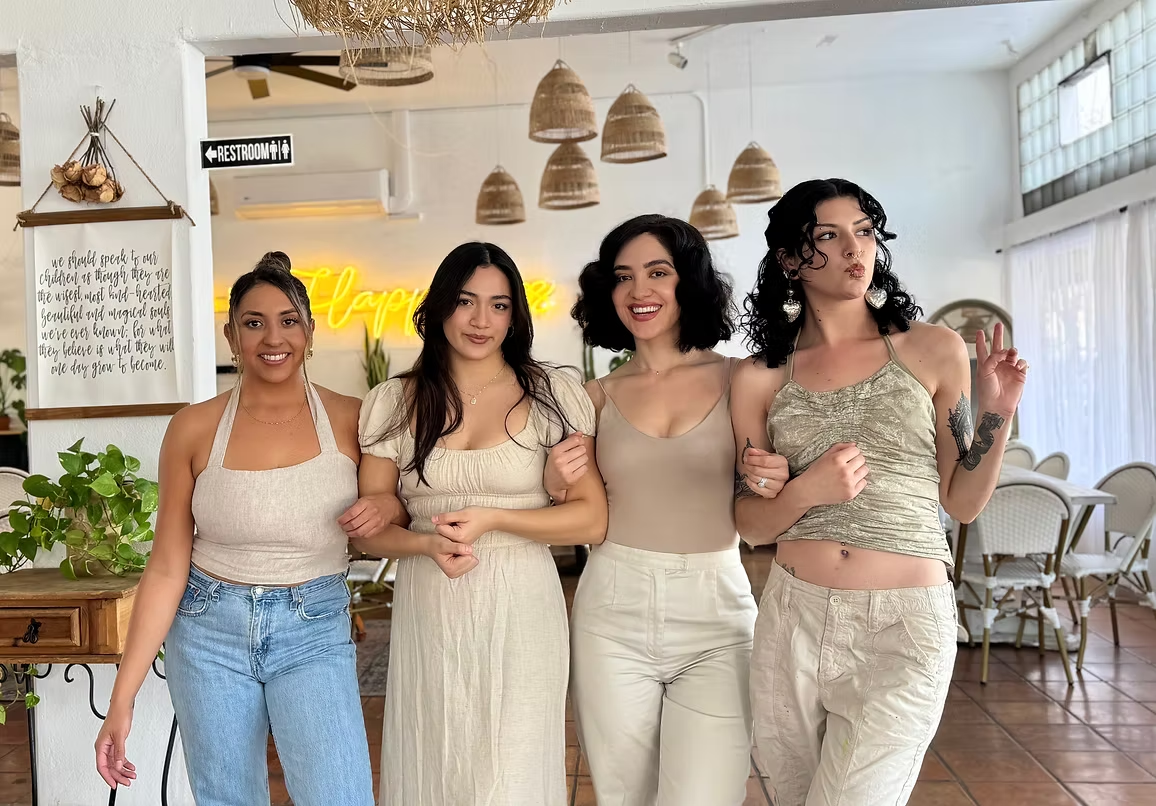 Four women arm in arm, smiling, in a bright, open-air cafe. Beige and white tones.