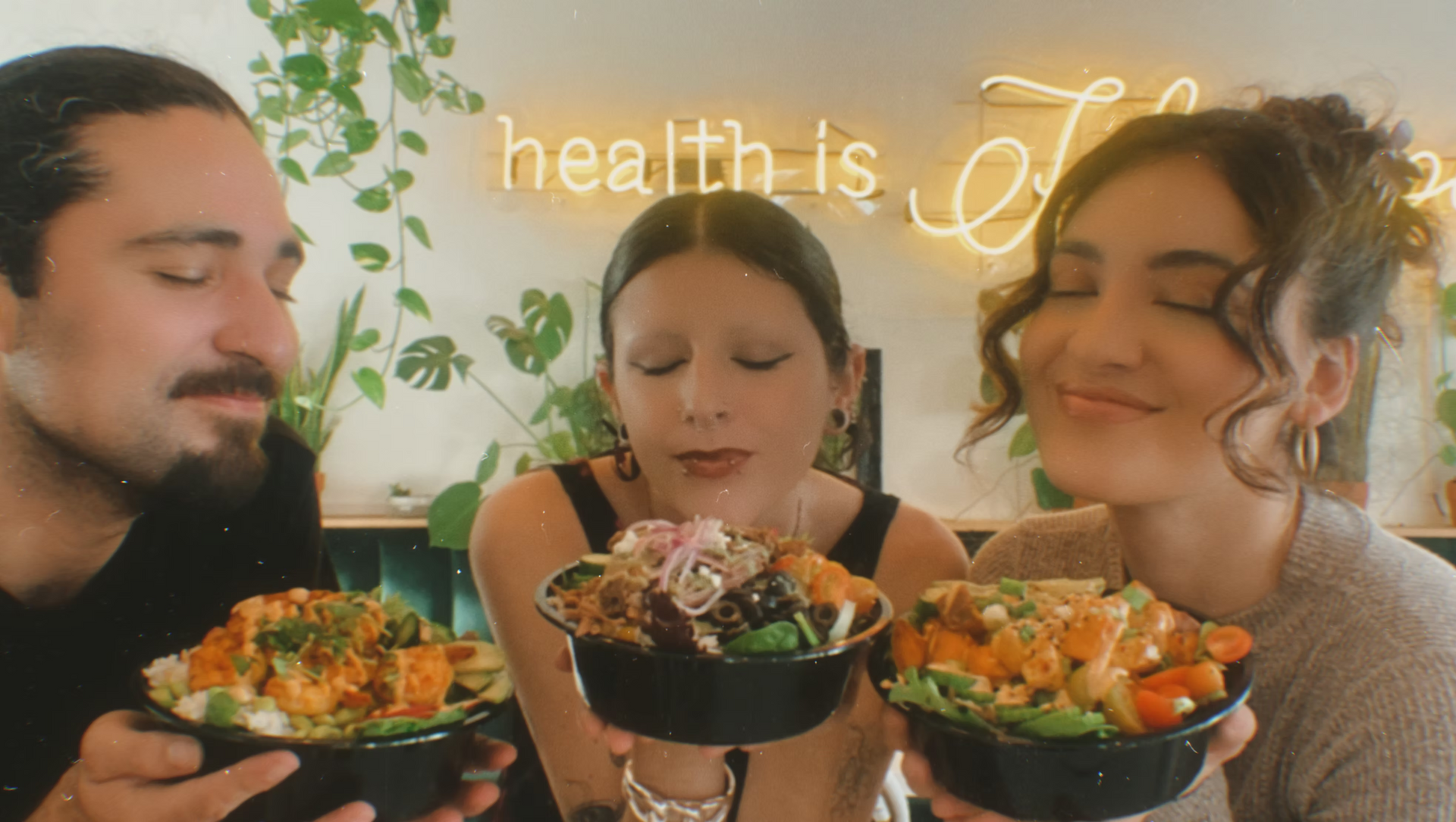 Three people smiling, holding and smelling bowls of food in a restaurant with neon sign saying