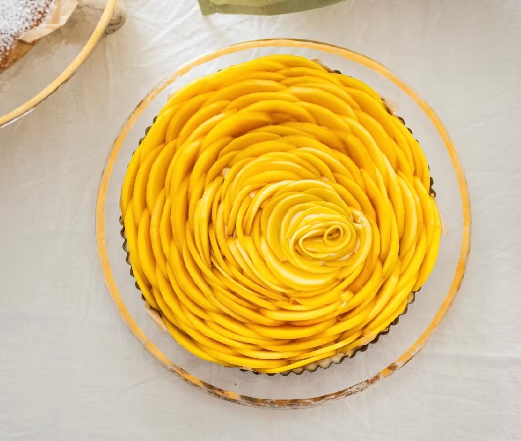 Mango tart with yellow mango slices arranged in a spiral pattern on a clear glass plate.