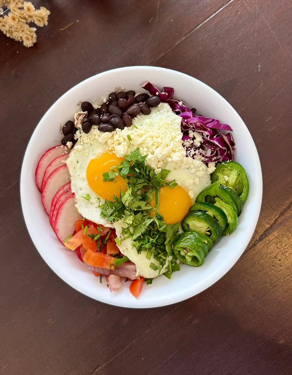 Breakfast bowl: fried eggs, black beans, red cabbage, radishes, jalapeños, pico de gallo, and cilantro in a white bowl.