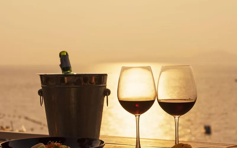 Two glasses of wine and a bucket of wine are on a table with a view of the ocean.