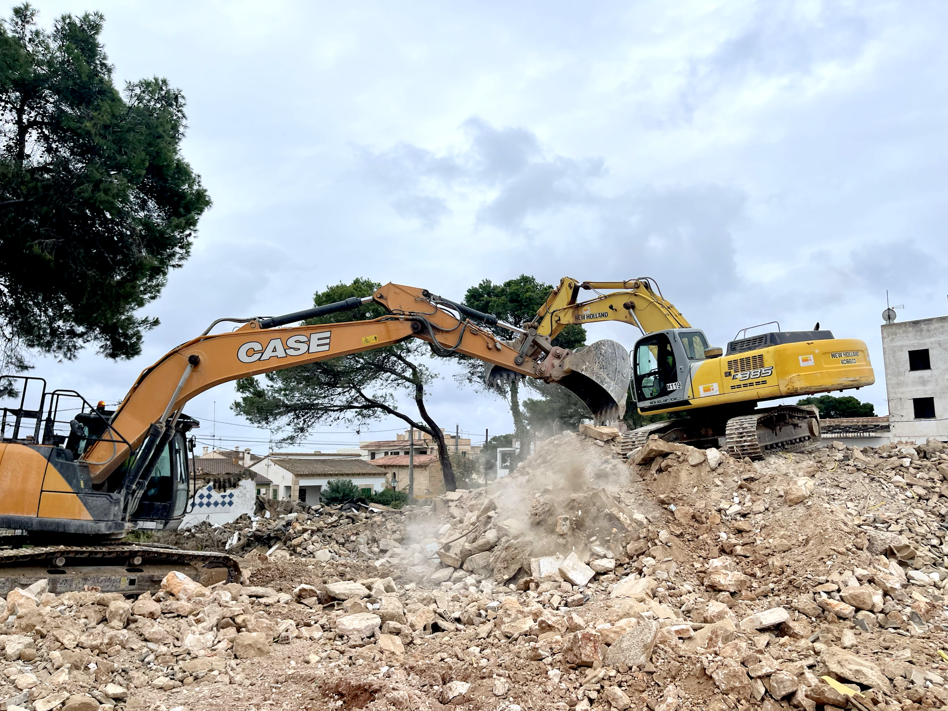 Dos excavadoras de construcción demoliendo un edificio en un día nublado, levantando polvo.