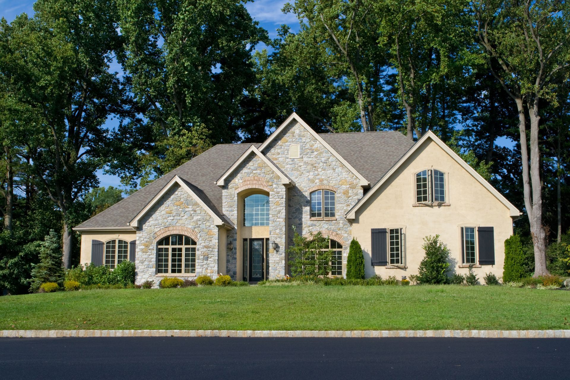 Stone and tan two-story house with black shutters, set in a green yard, with trees in the background. Stone and tan two-story house with black shutters, set in a green yard, with trees in the background.