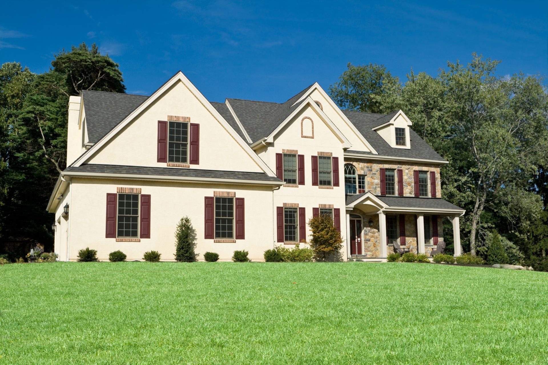 Two-story house with tan siding, burgundy shutters, and stone facade, set against green lawn and blue sky. Two-story house with tan siding, burgundy shutters, and stone facade, set against green lawn and blue sky.