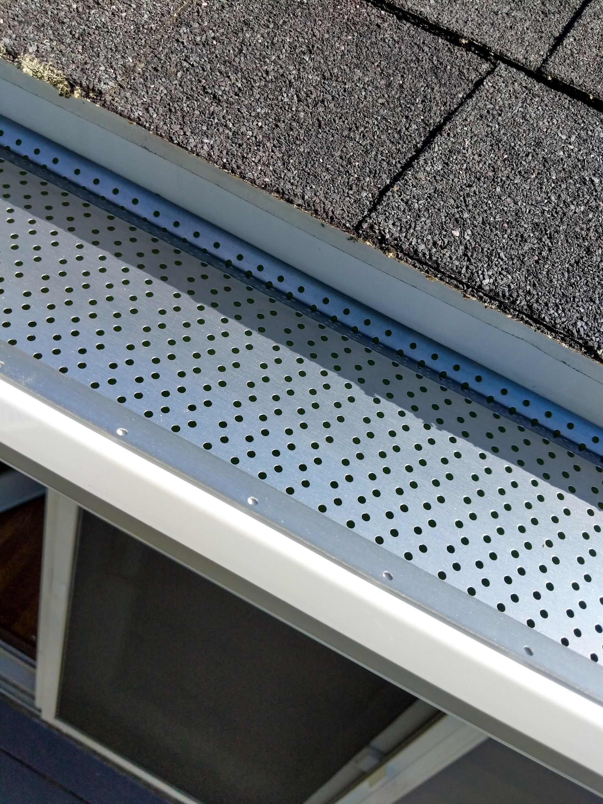 Close-up of house gutter with mesh screen, installed along a shingled roofline. Close-up of house gutter with mesh screen, installed along a shingled roofline.