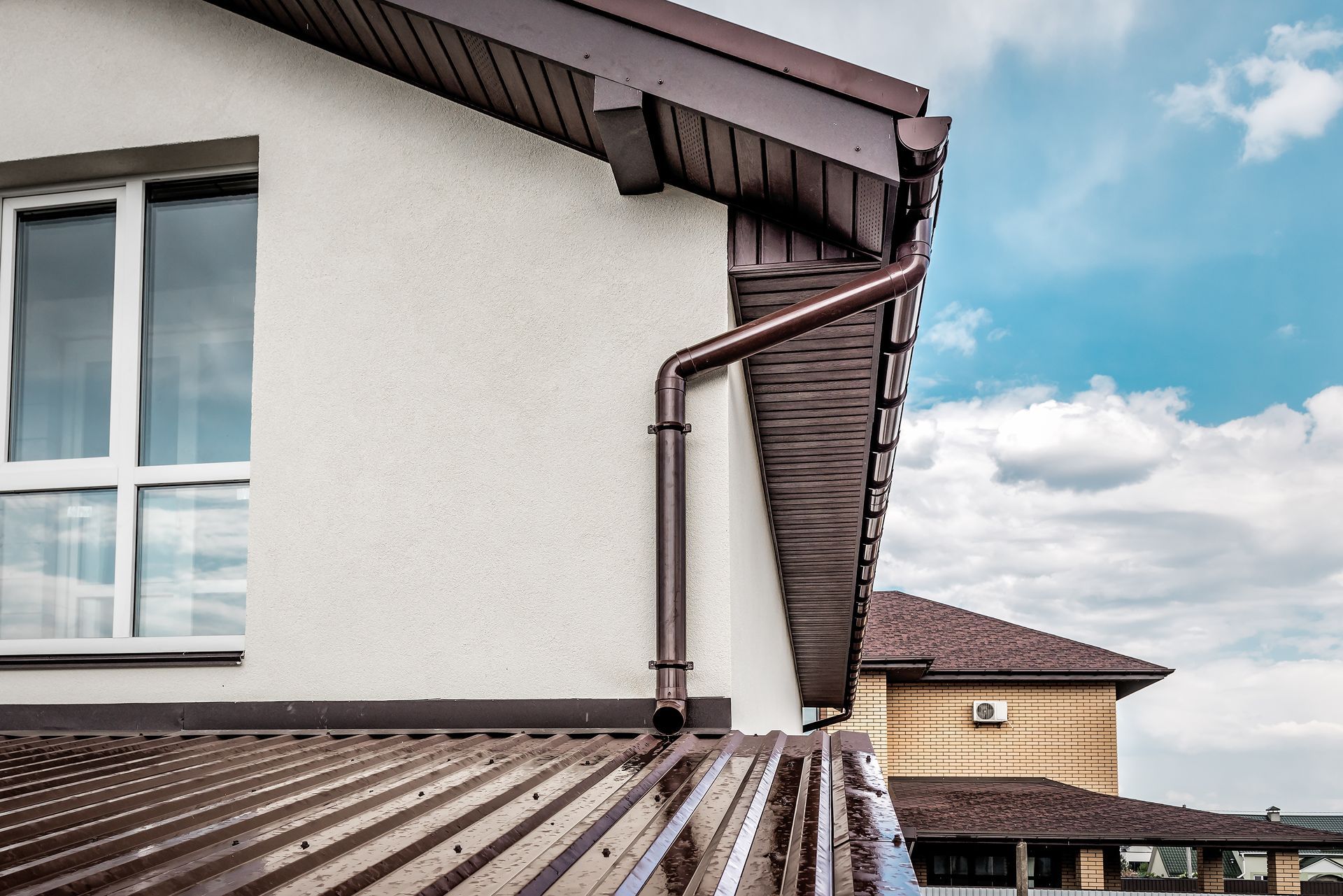 Brown gutter and downspout on a white stucco building with a window and a rooftop, blue sky. Brown gutter and downspout on a white stucco building with a window and a rooftop, blue sky.