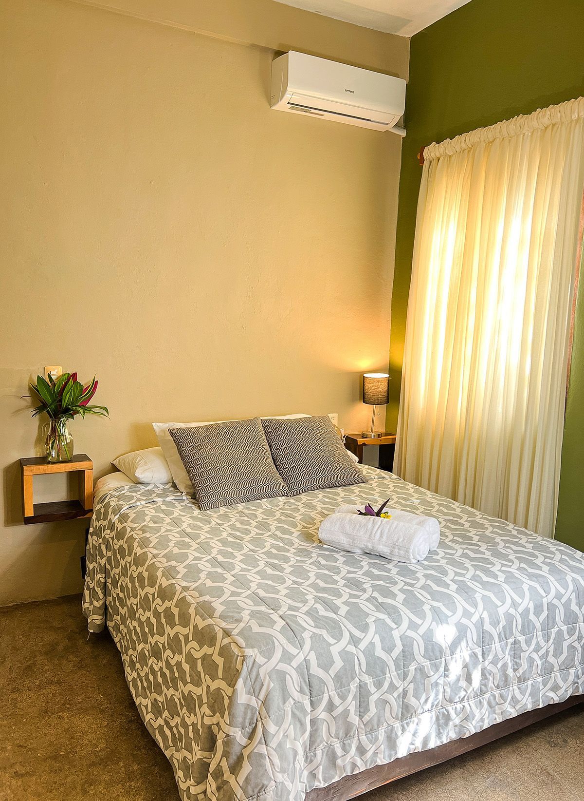 A hotel room with a bed , nightstand , lamp and air conditioner.