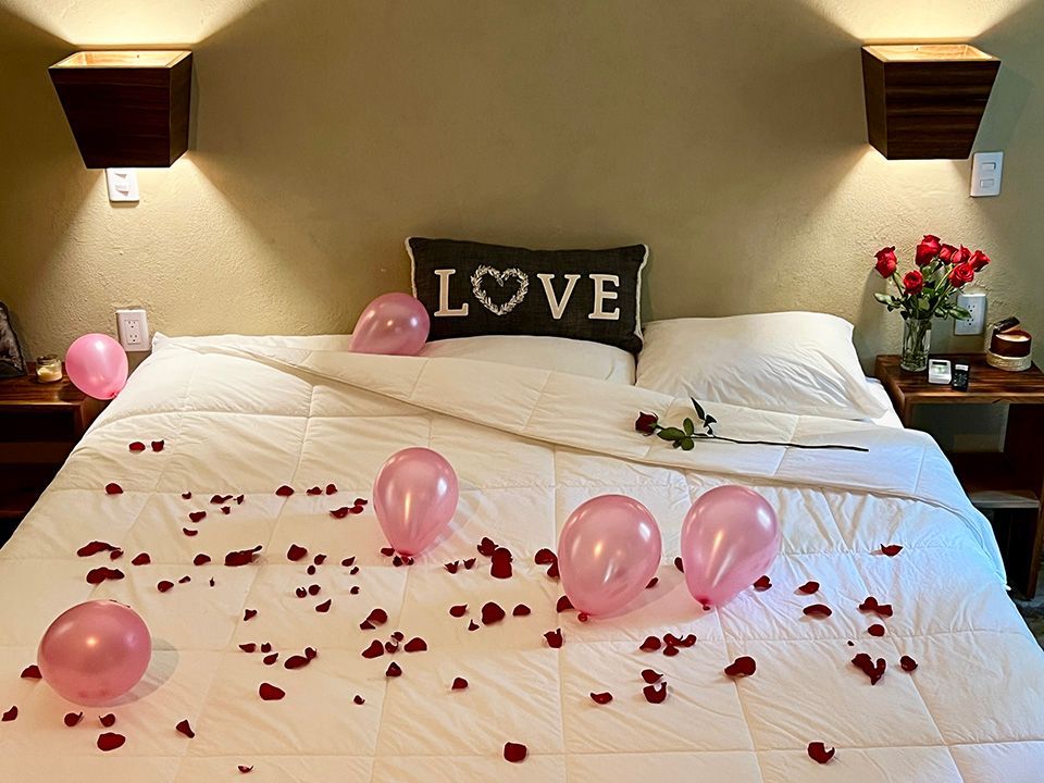 A bed with a pillow that says love on it and some balloons and rose petals.