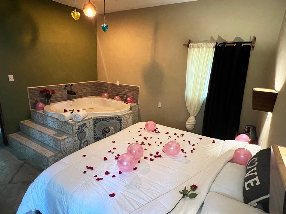 A hotel suite with a bed and a jacuzzi surrounded by balloons and rose petals.
