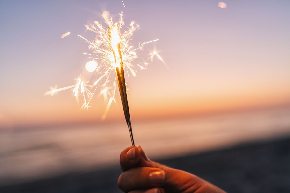 A hand holding a glowing sparkler on Carolina Beach at sunset, celebrating the festive coastal vibes of winter by the ocean.