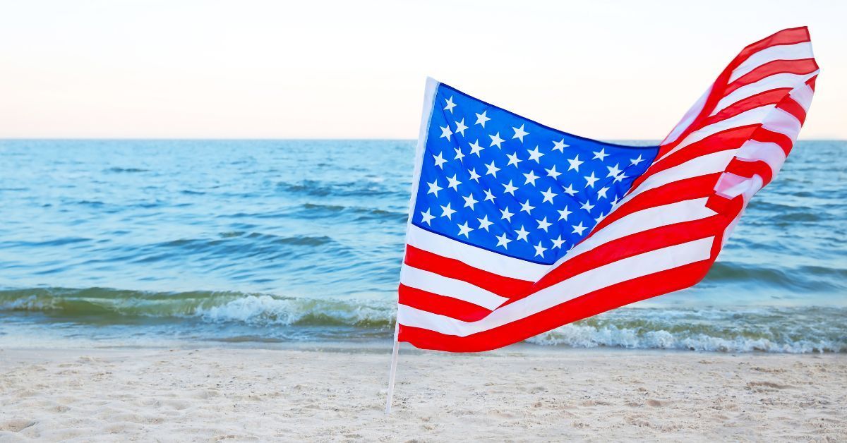 A vibrant American flag anchored in the sand, gently blowing in the breeze with the ocean waves in the background. This image captures a patriotic, serene beach setting perfect for military families and visitors.
