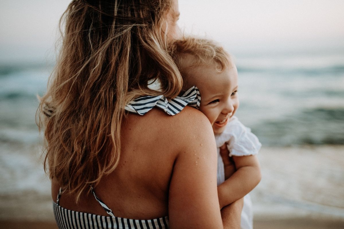 A smiling mother holds her laughing baby at the beach near Carolina Beach Inn. The ocean waves and soft light create a warm and serene moment.