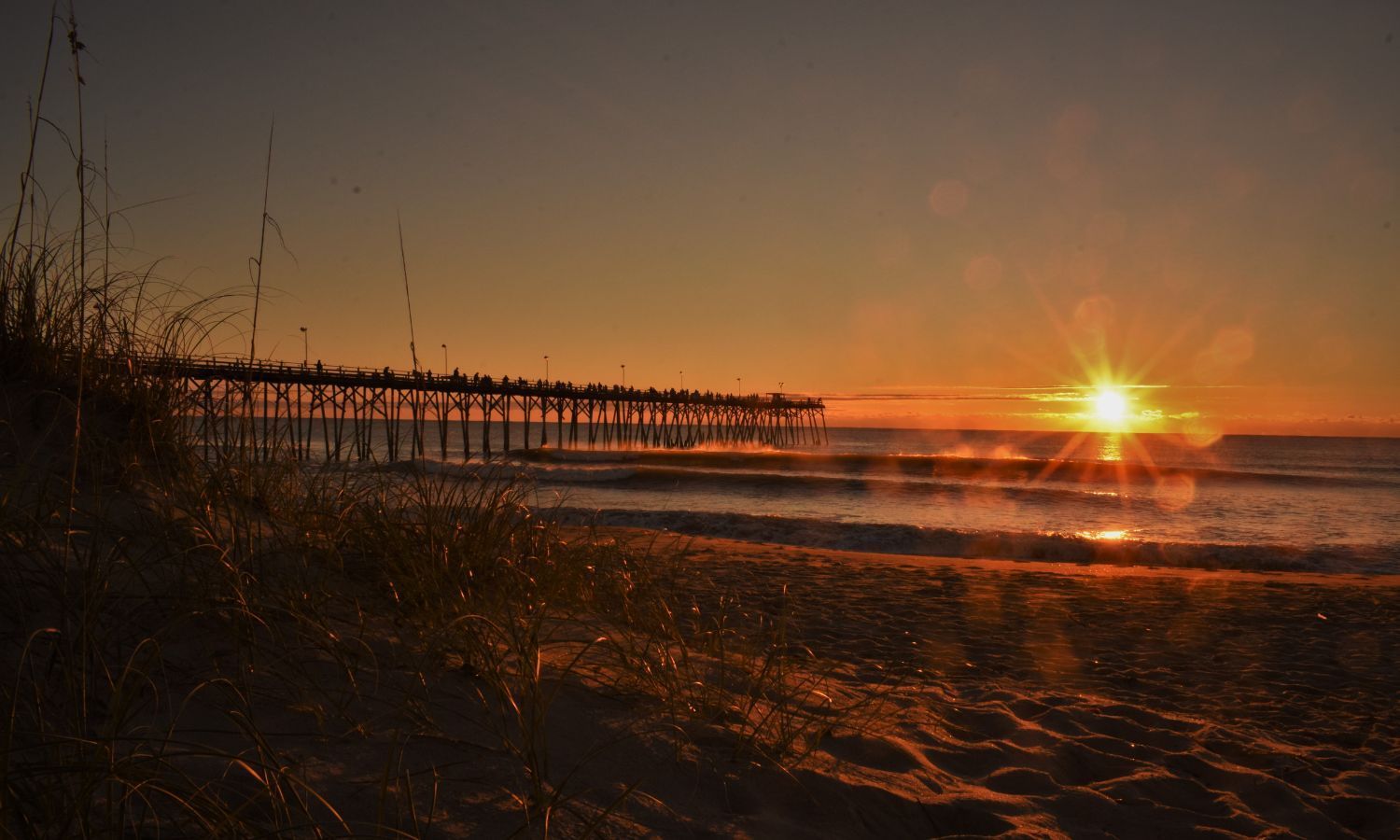 Sunrise at Kure Beach pier with warm orange hues reflecting off the ocean waves, grasses