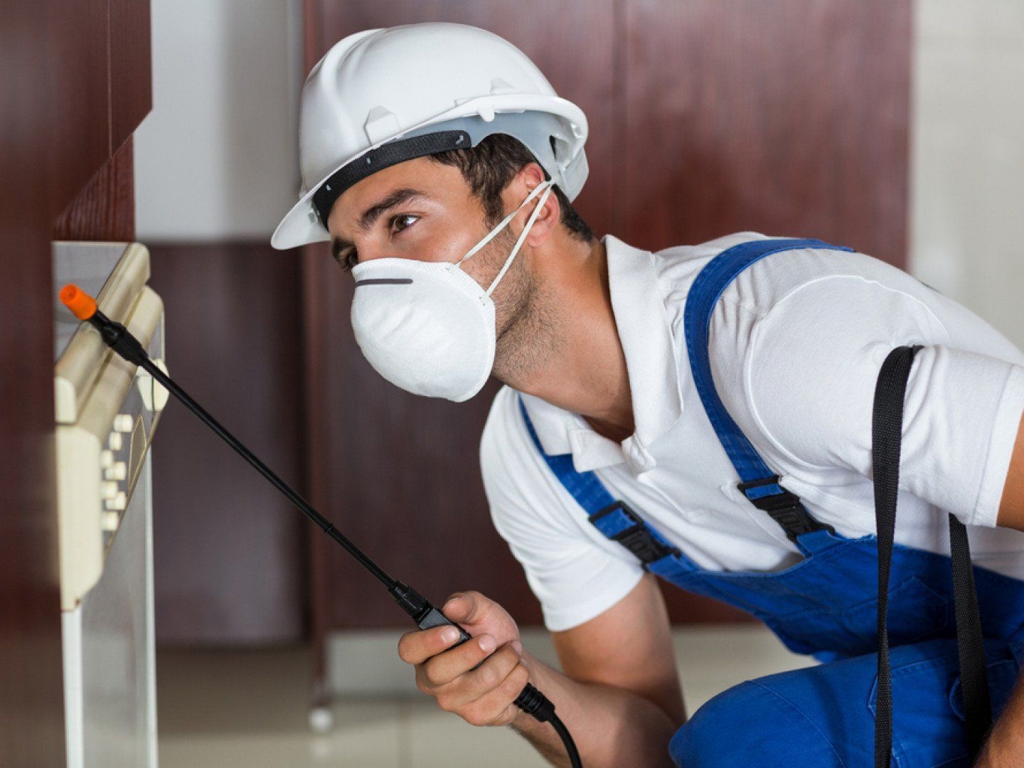Pest control worker spraying insecticide near a cabinet, wearing a respirator, hard hat, and blue overalls.