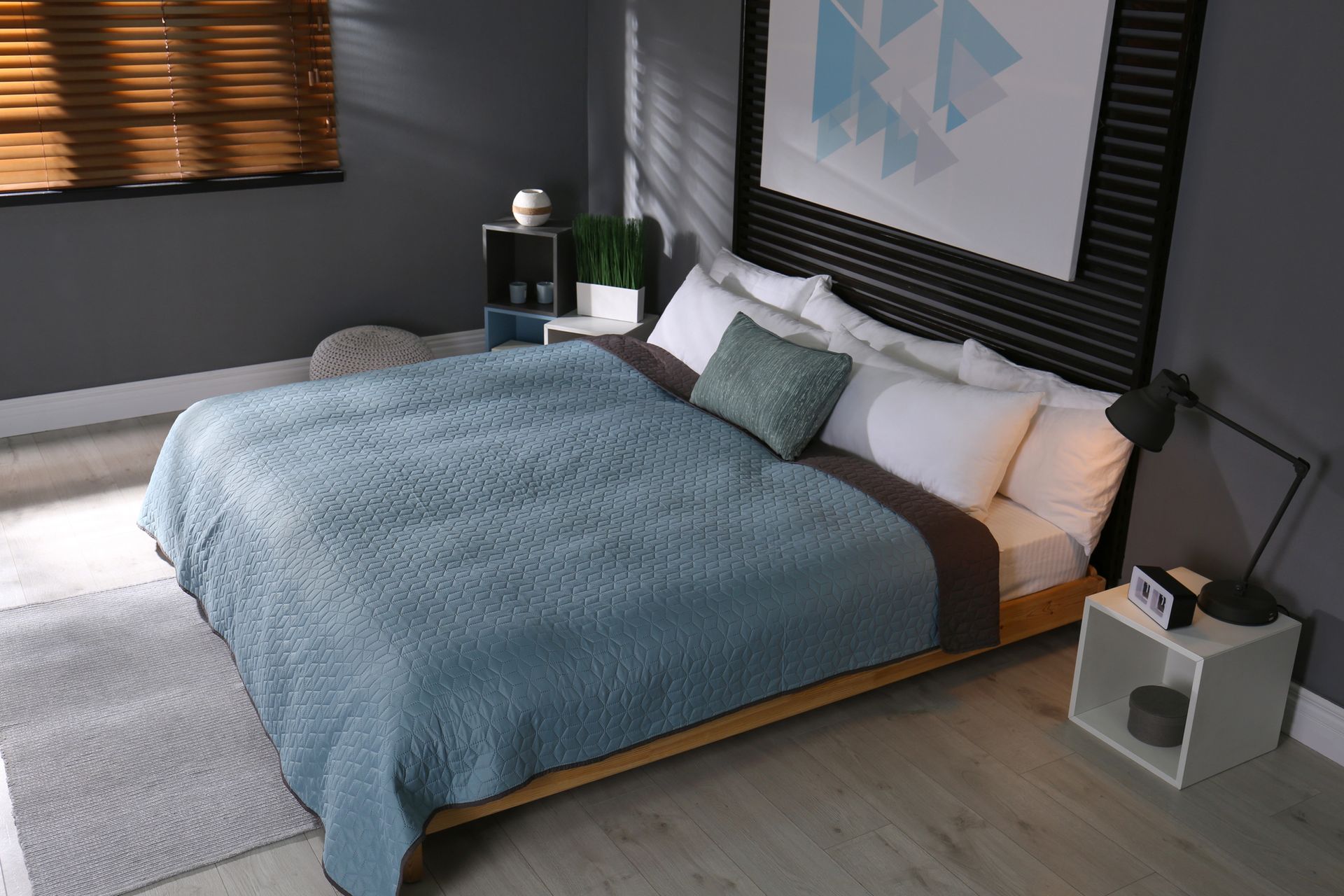 Dormitorio moderno con colcha azul, almohadas blancas y persianas oscuras.